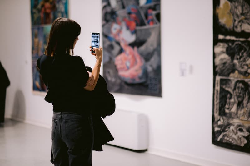 Devojka u crnoj majici fotografiše telefonom strip tapiseriju za izložbi Heroji svakodnevice